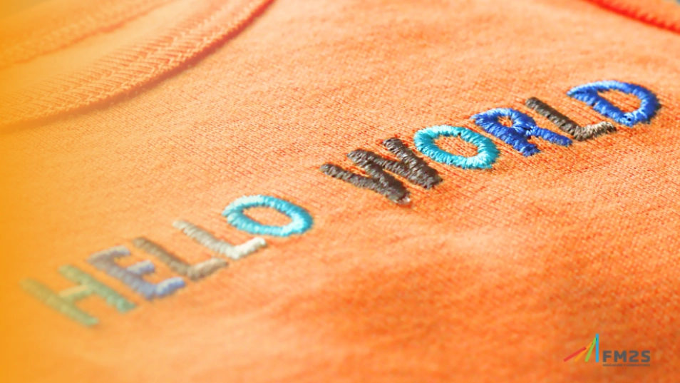 Degradê laranja à esquerda. Frase "Hello world" bordada em tecido laranja. Logo da FM2S no canto inferior direito.
